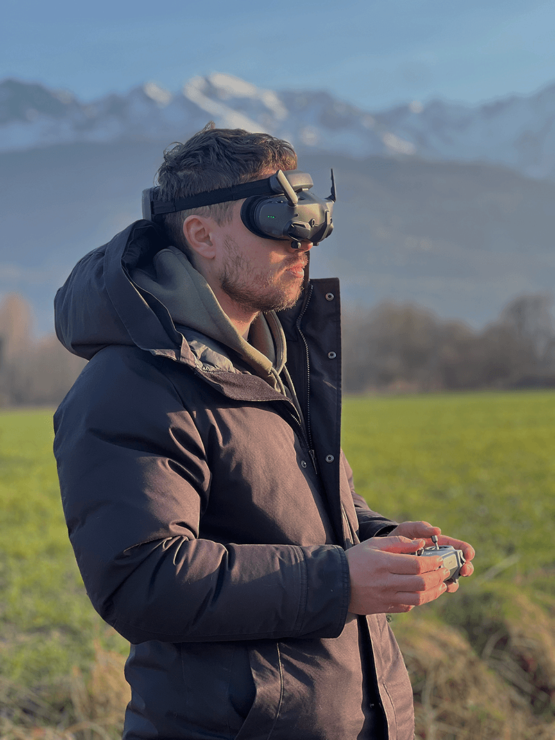 Homme portant un casque FPV et tenant une radiocommande debout dans un champ avec des montagnes en arrière-plan.