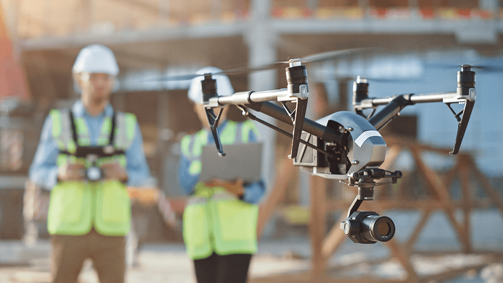 Drone en vol avec deux techniciens en gilet de sécurité et casque blanc sur un chantier.