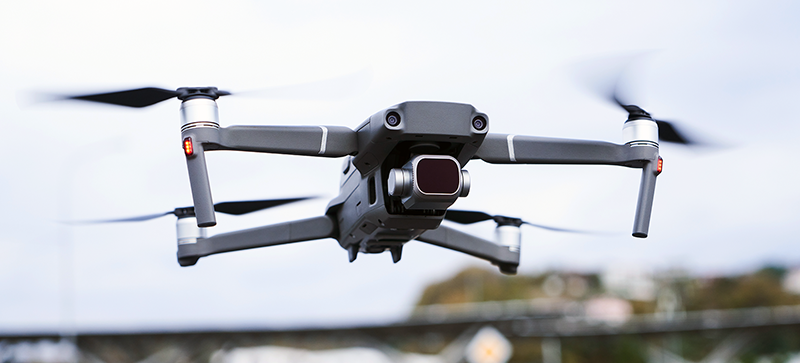 Drone gris volant avec caméra frontale en extérieur, fond flou avec arbres et ciel clair.