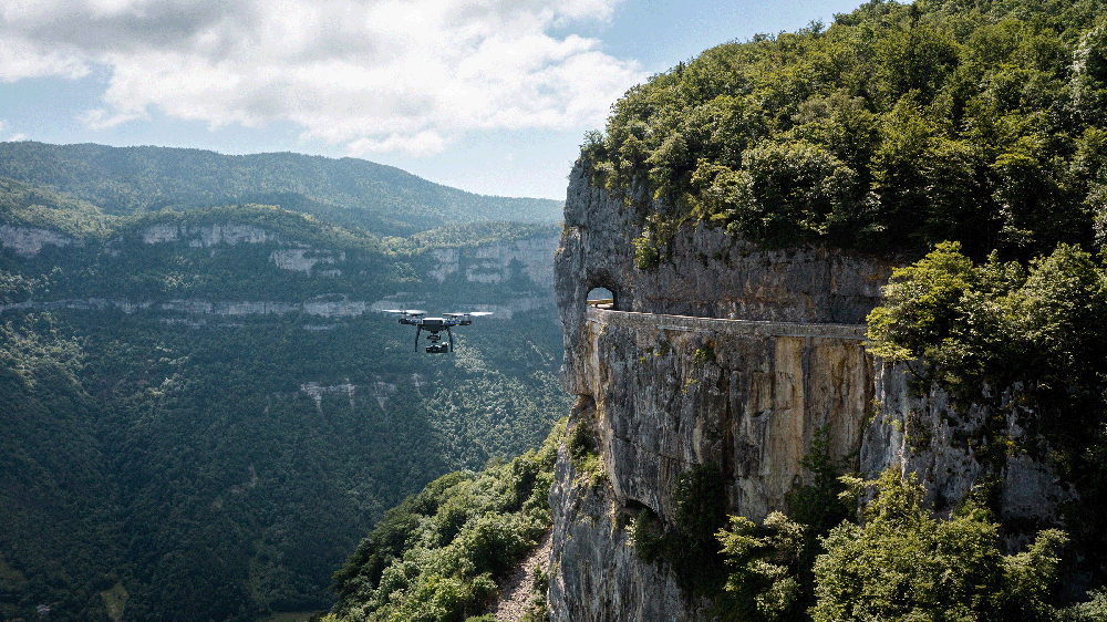 Un drone vole devant une falaise boisée avec une route taillée et une arche dans la roche.