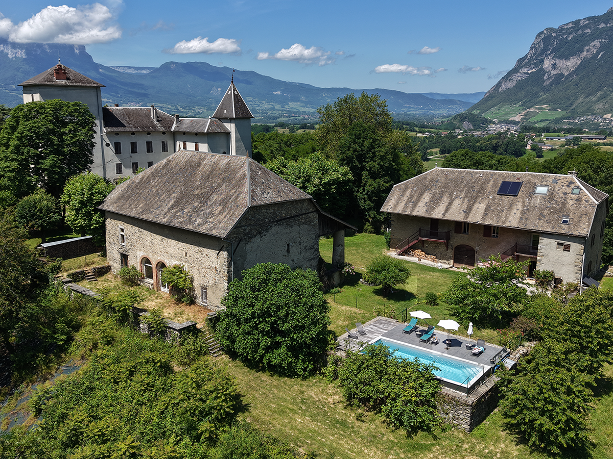 Vue aérienne d'un chalet et piscine entourés d'arbres avec montagnes en arrière-plan par beau temps.