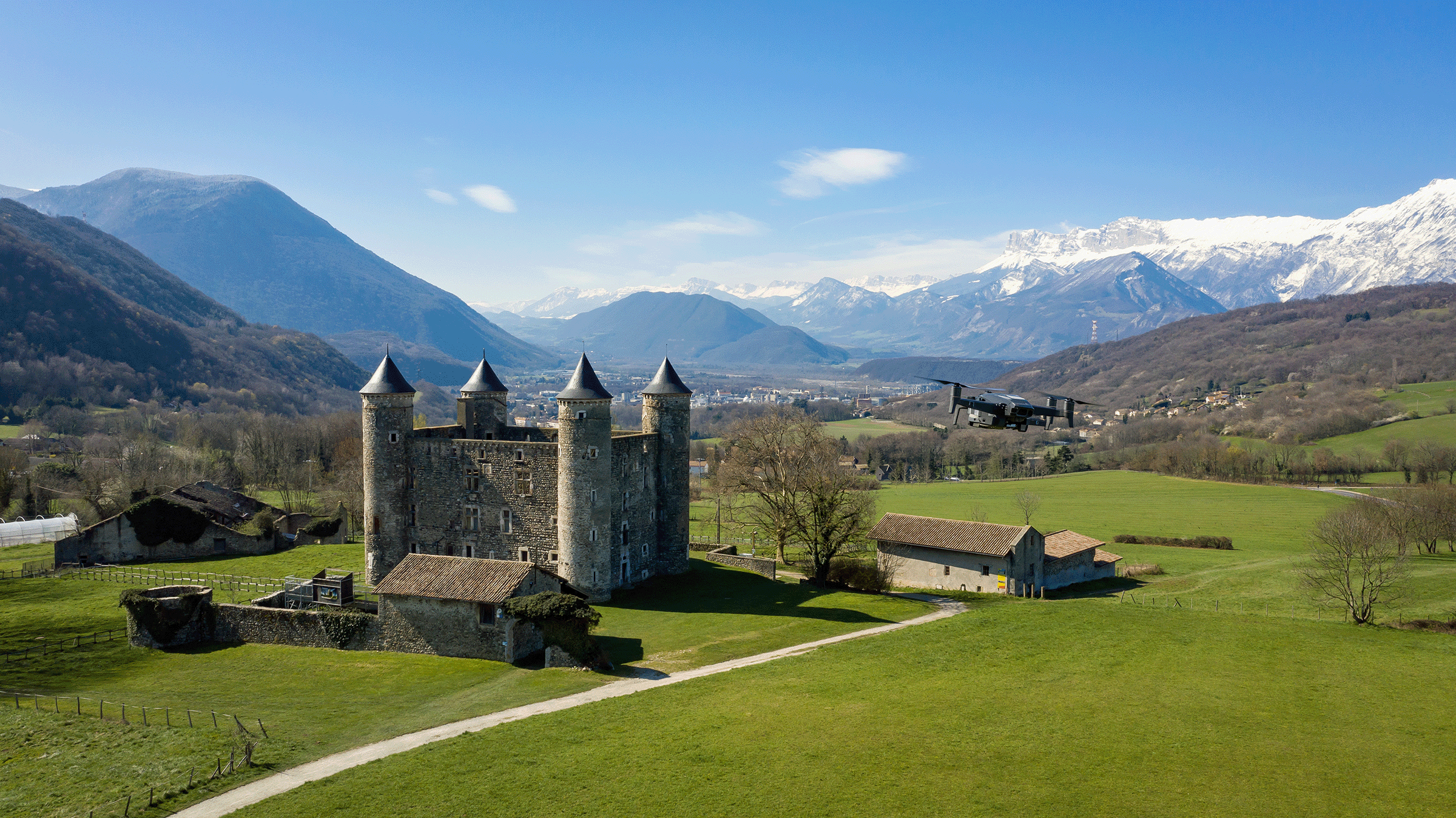 Un château médiéval dans la campagne, montagnes enneigées en arrière-plan et un drone volant à droite.