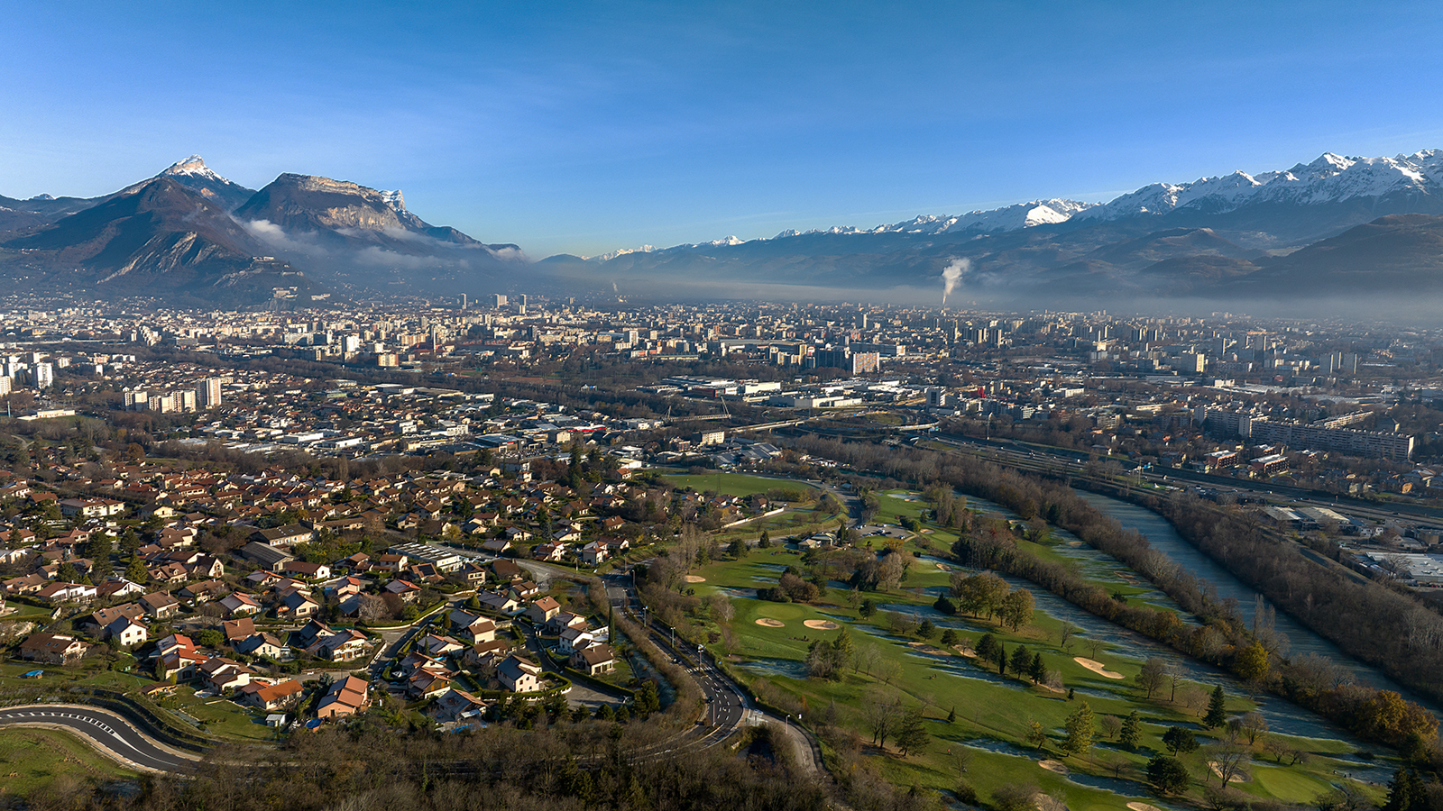 Vue aérienne d'une ville entourée de maisons, rivière et montagnes enneigées par ciel clair.