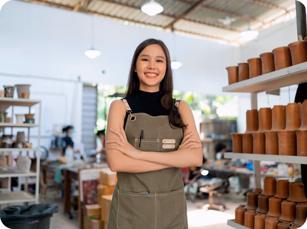 Smiling woman wearing an apron with arms crossed standing in a pottery workshop with shelves of clay pots.