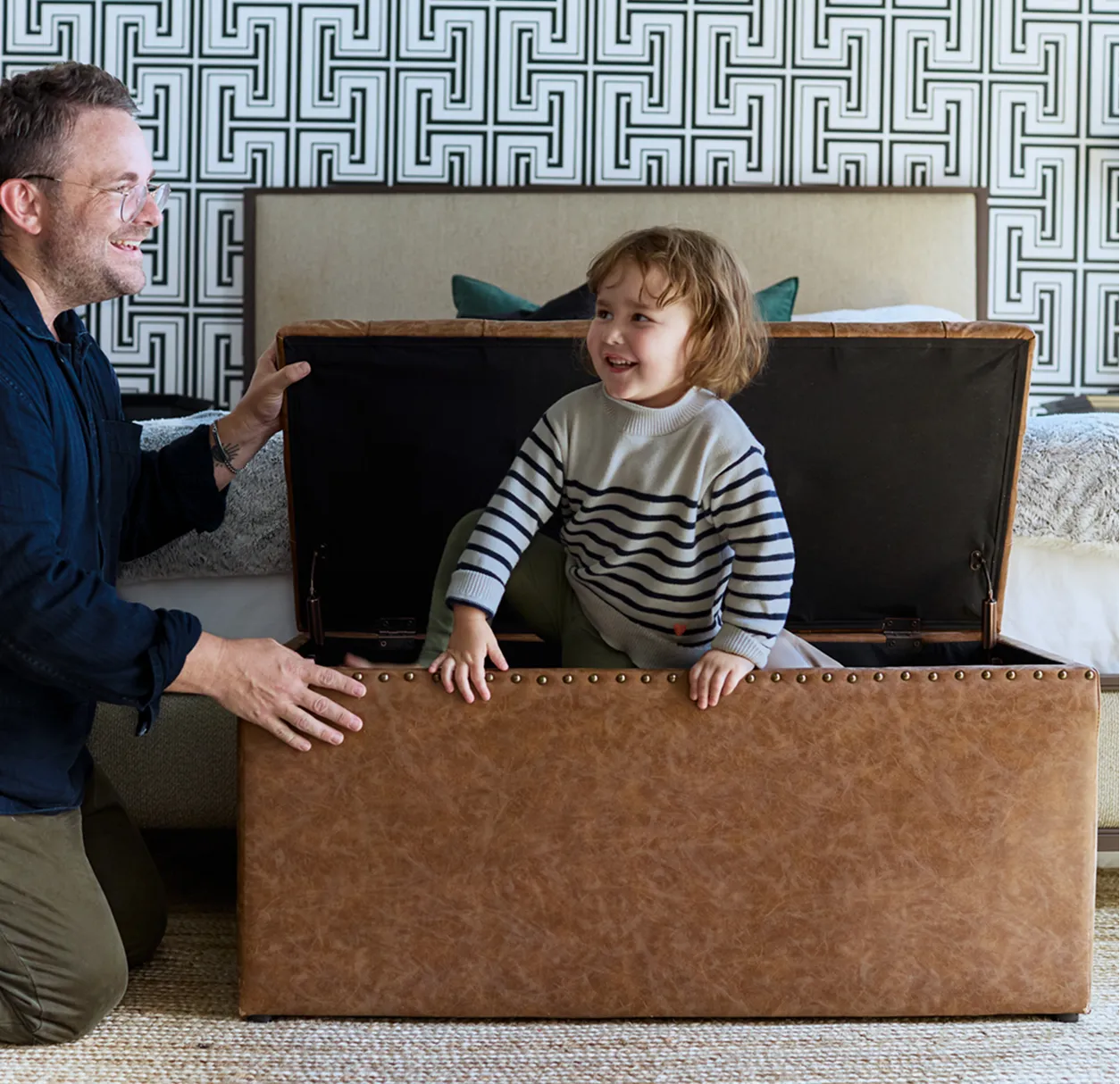 Smiling man opens a large brown storage chest while a young child sits inside it in a patterned bedroom.