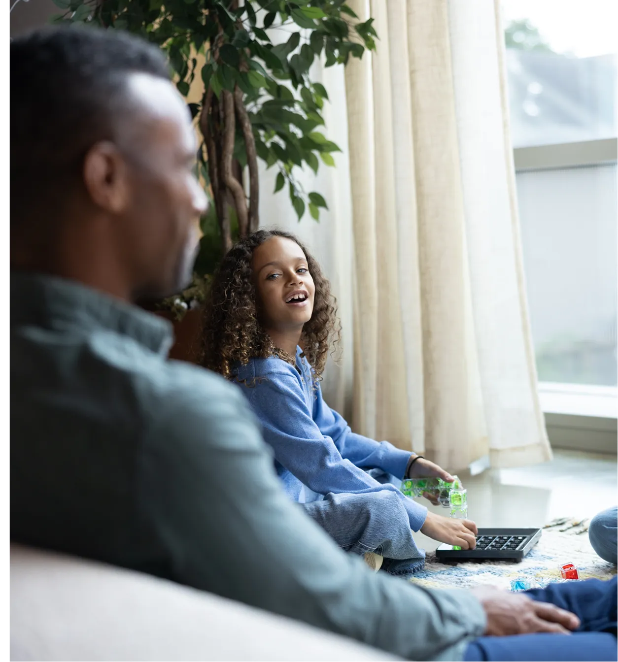 Smiling girl with curly hair playing a board game on the floor near a man sitting on a sofa.