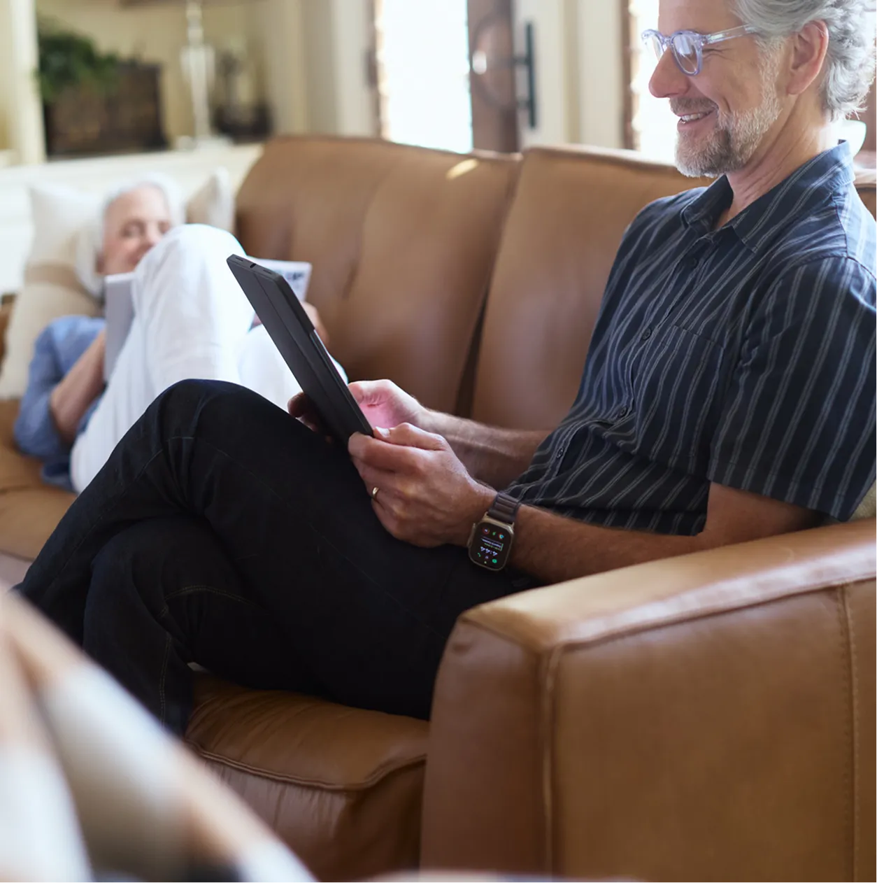 Smiling elderly man sitting on a brown leather sofa using a tablet while an elderly woman reclines in the background reading a book.