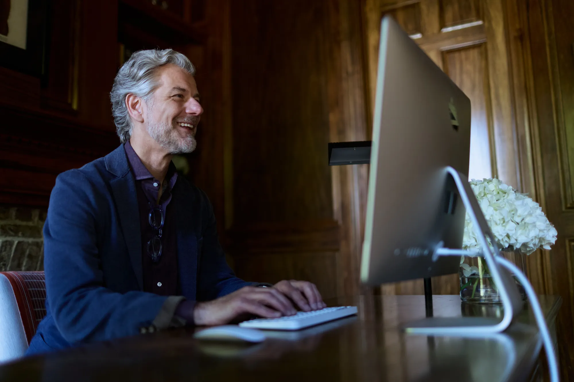 Smiling middle-aged man with gray hair and beard typing on a desktop computer in a wood-paneled room.