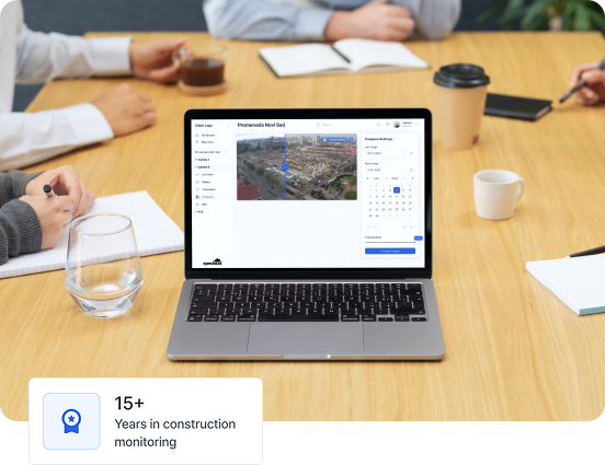 Laptop on a wooden table showing construction monitoring software with aerial images, surrounded by people with notebooks and coffee cups.