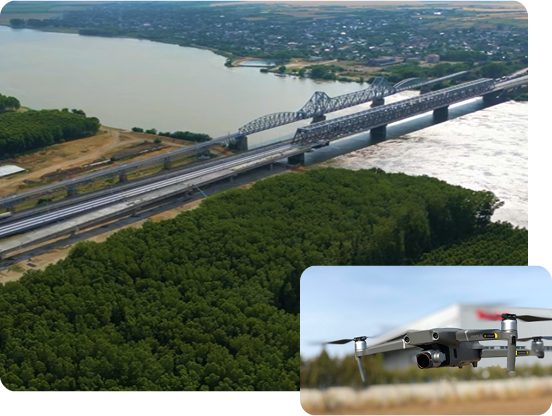 Aerial view of a long bridge over a wide river with dense green forest in the foreground and a city in the background, inset shows a close-up of a gray drone flying outdoors.