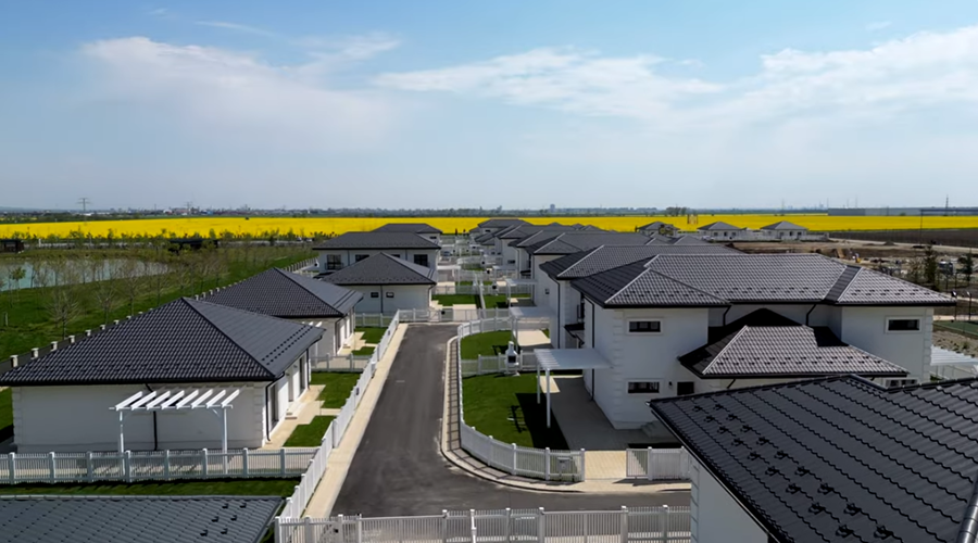 Aerial view of a residential neighborhood with modern houses featuring dark roofs, a paved road, green lawns, and yellow fields in the distance.