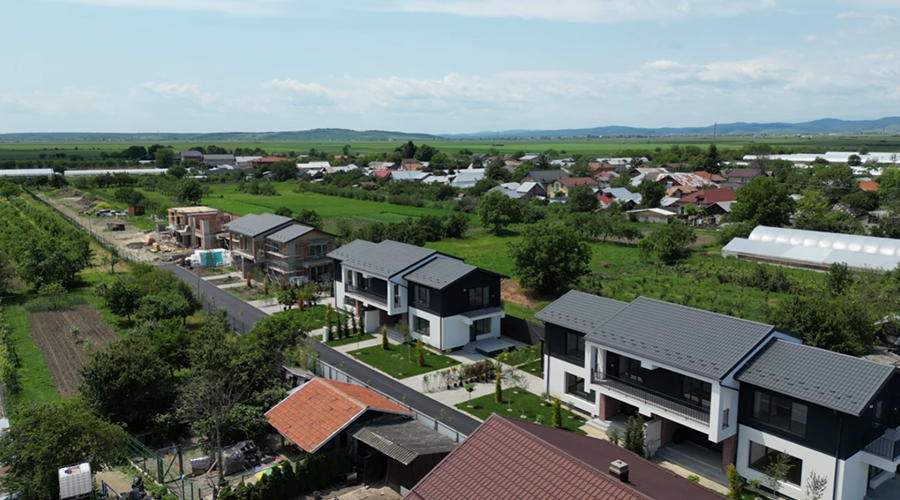 Aerial view of a residential neighborhood with modern two-story houses, green fields, and distant hills under a partly cloudy sky.
