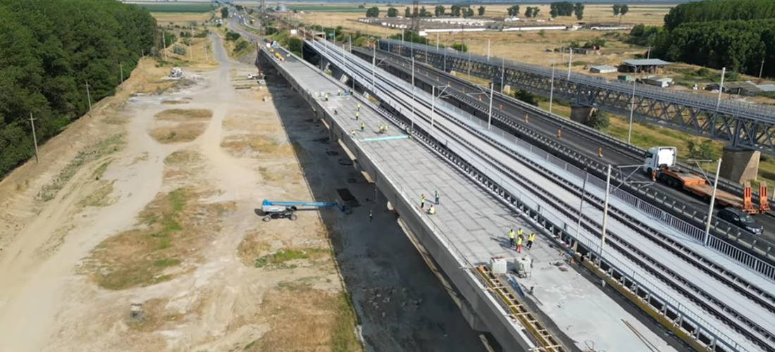 Aerial view of a highway bridge under construction next to an operational bridge with vehicles, surrounded by fields and trees.