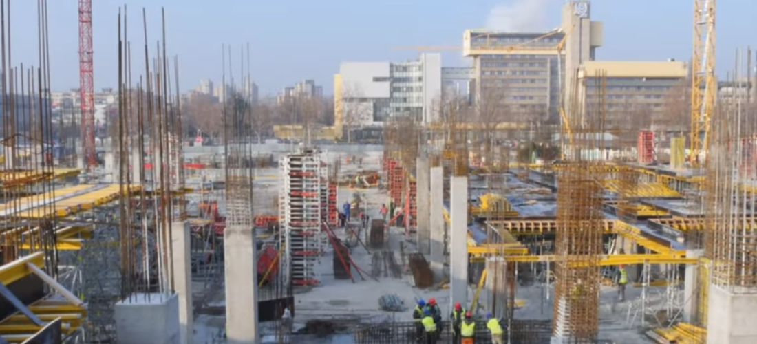 Construction site with workers wearing helmets and safety vests assembling concrete and steel structures amid cranes and urban buildings in the background.