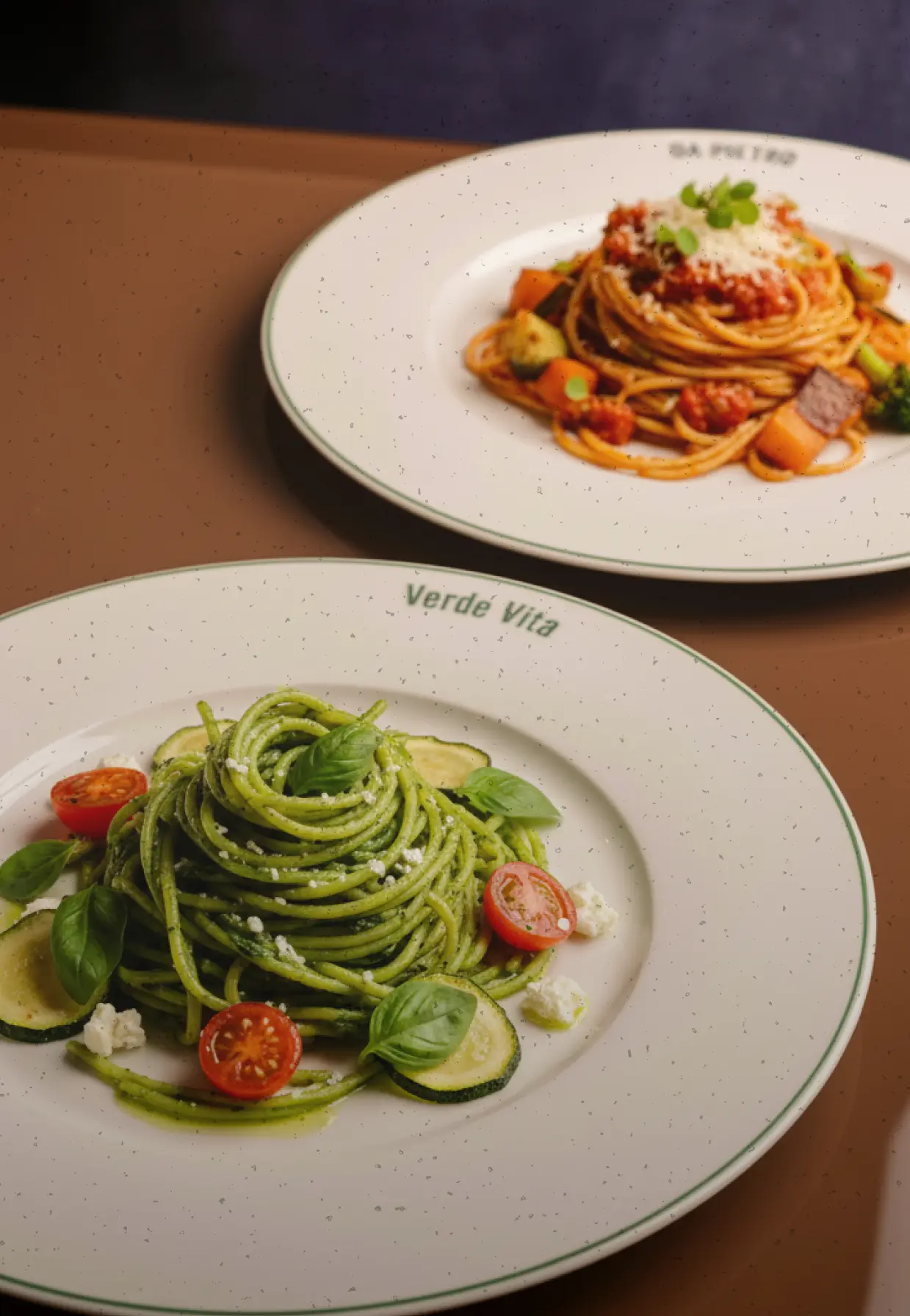 Two plates of spaghetti pasta, one with pesto sauce, cherry tomatoes, zucchini, basil, and ricotta cheese, the other with red sauce, grated cheese, and mixed vegetables.