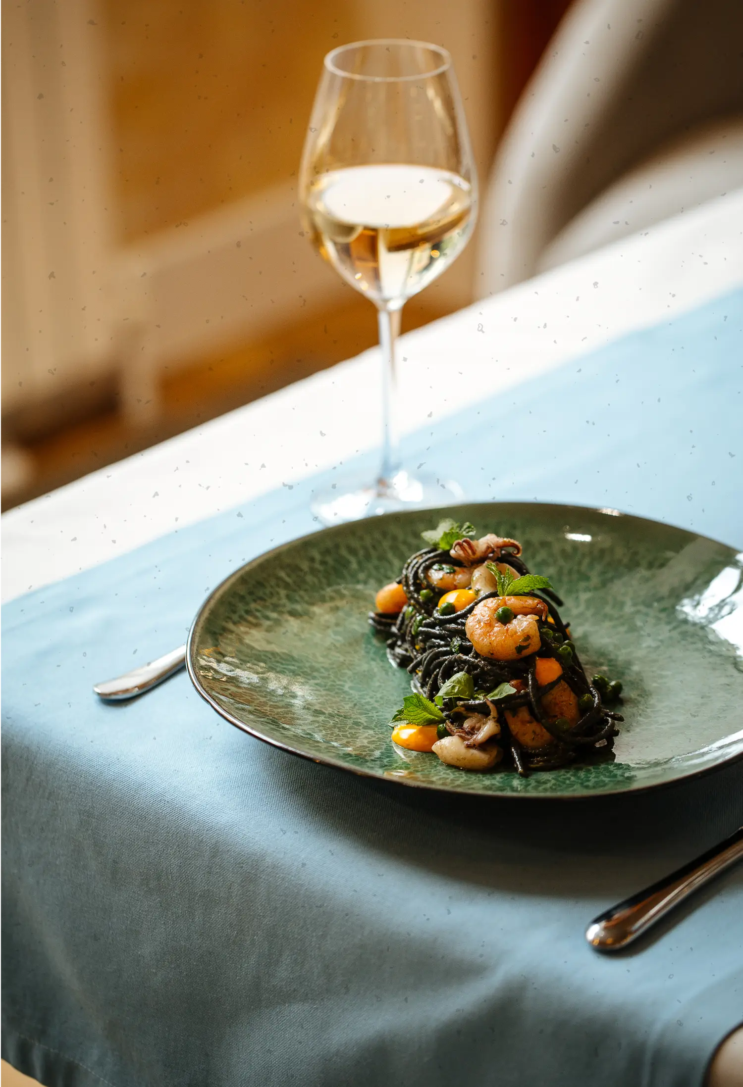 Green plate with squid ink pasta, shrimp, and herbs, accompanied by a glass of white wine on a table with a blue tablecloth.