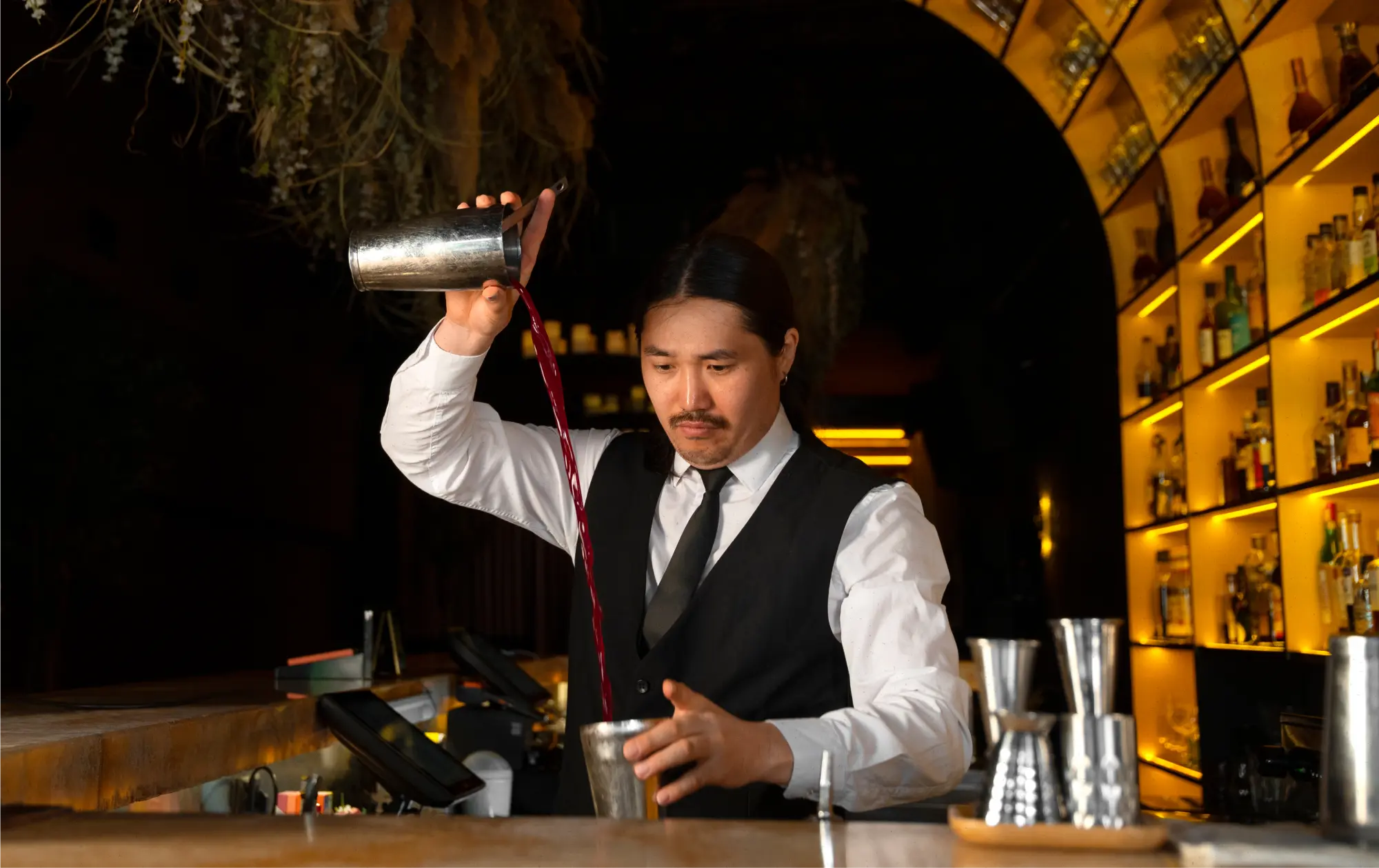 Bartender in a white shirt and black vest pouring a red liquid from one shaker to another behind a bar with illuminated shelves of liquor bottles.