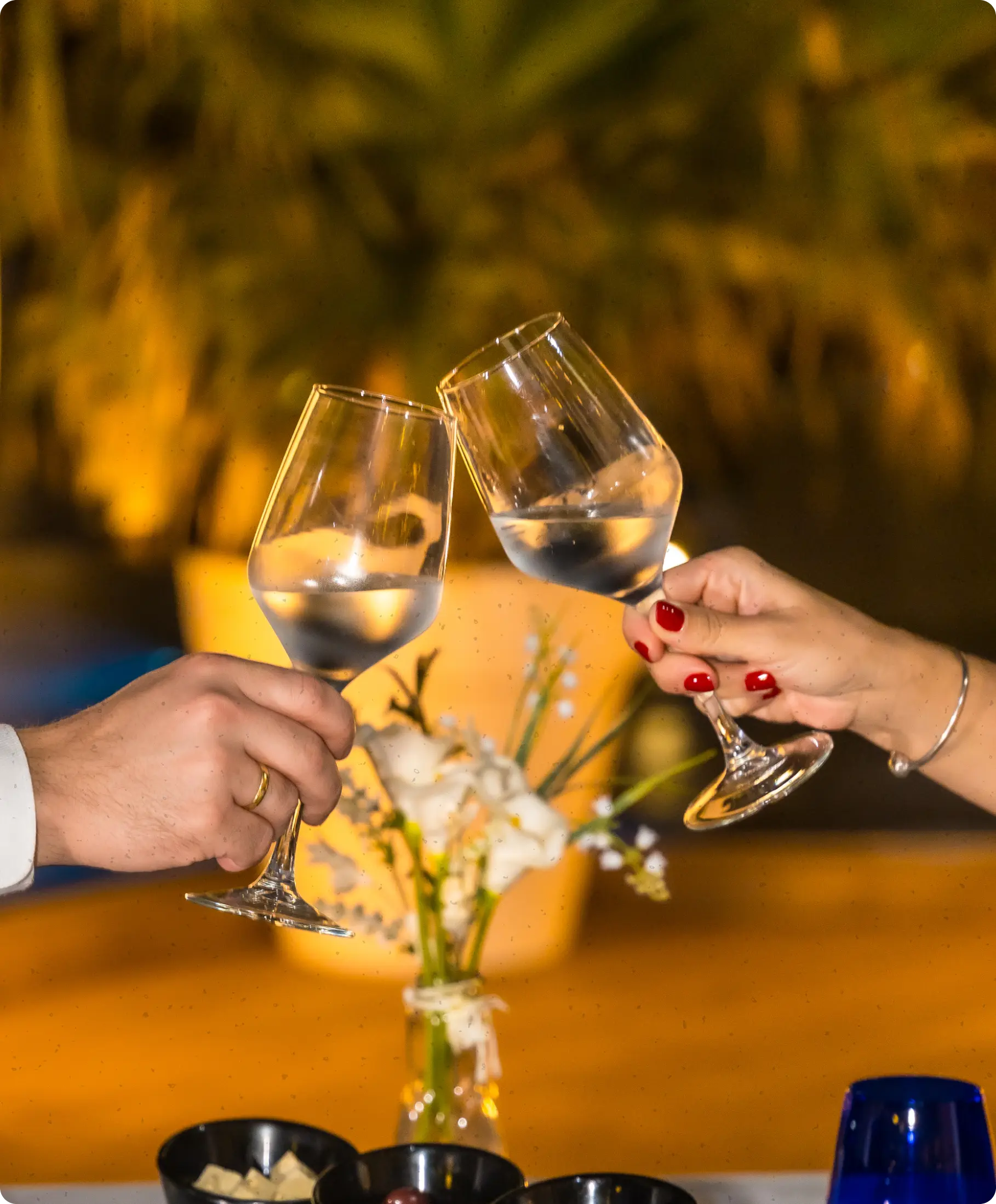 Two hands clinking wine glasses filled with a clear liquid during a toast, with a blurred flower vase and warm lighting in the background.