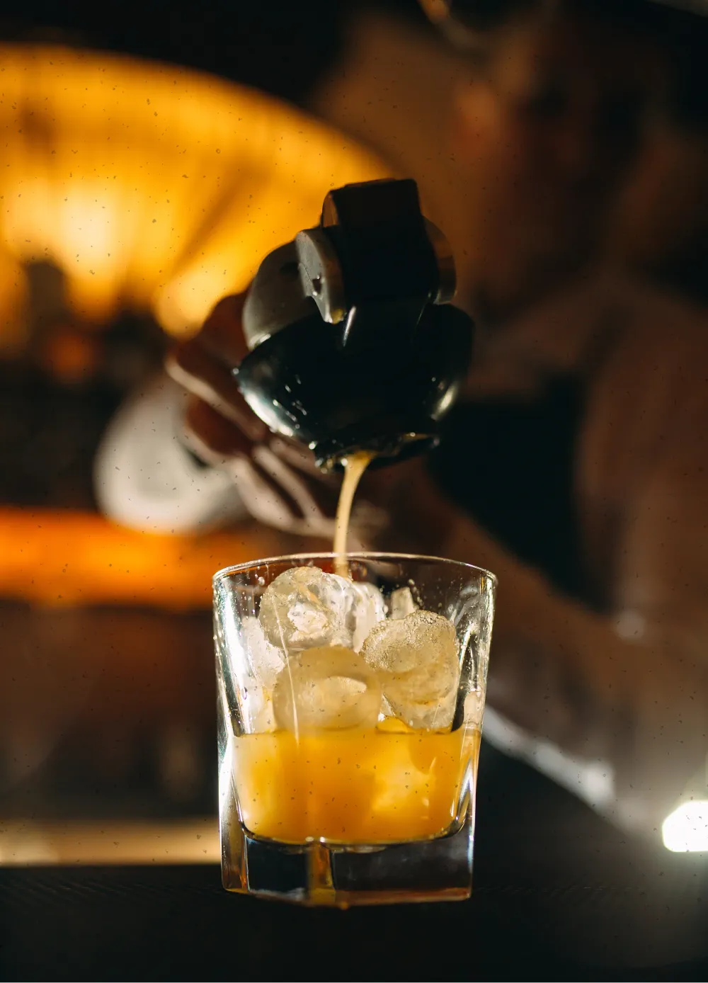 Close-up of a bartender pouring a yellow-orange cocktail over ice in a glass at a dimly lit bar.