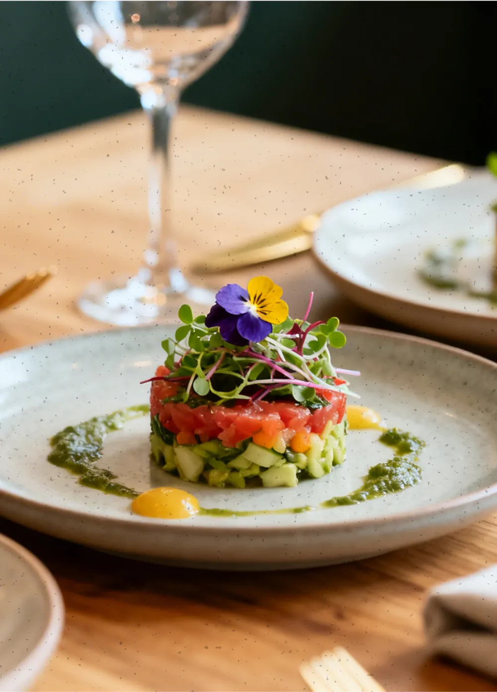 Plated gourmet vegetable tartare topped with microgreens and an edible purple and yellow flower, garnished with green herb sauce and yellow dollops on a light ceramic plate.