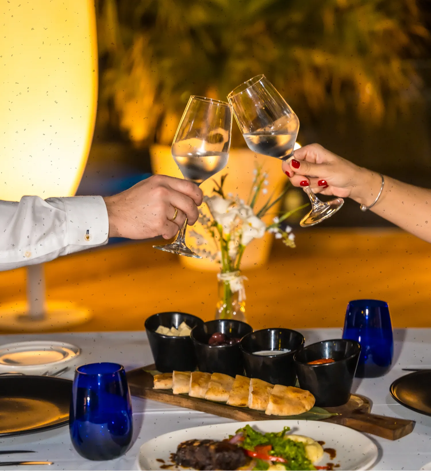 Two hands clinking wine glasses filled with clear liquid over a table set with bread, dips, and a plated meal in warm ambient lighting.