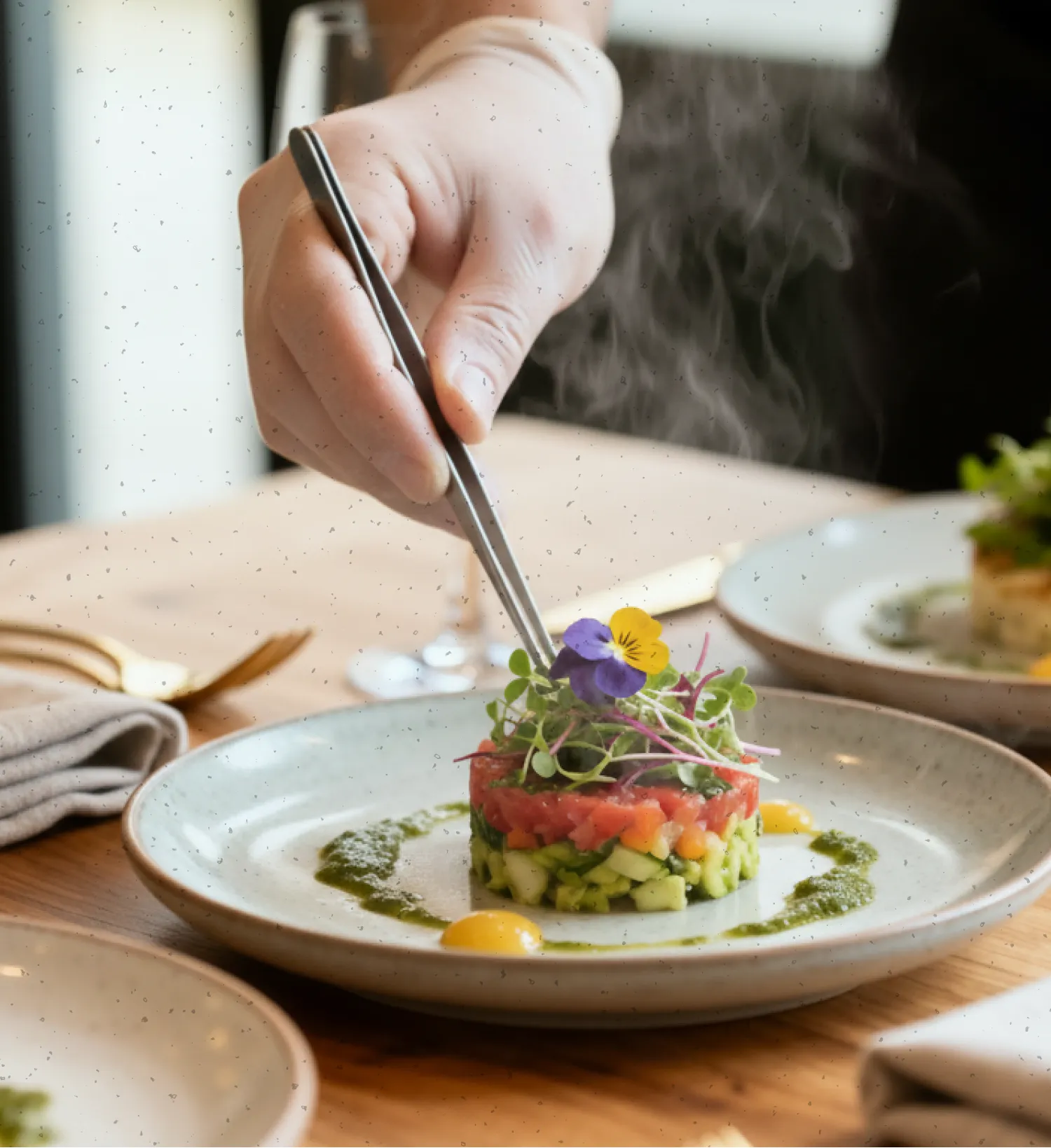 A chef's hand using tweezers to garnish a layered vegetable and herb dish with edible flowers on a white plate.