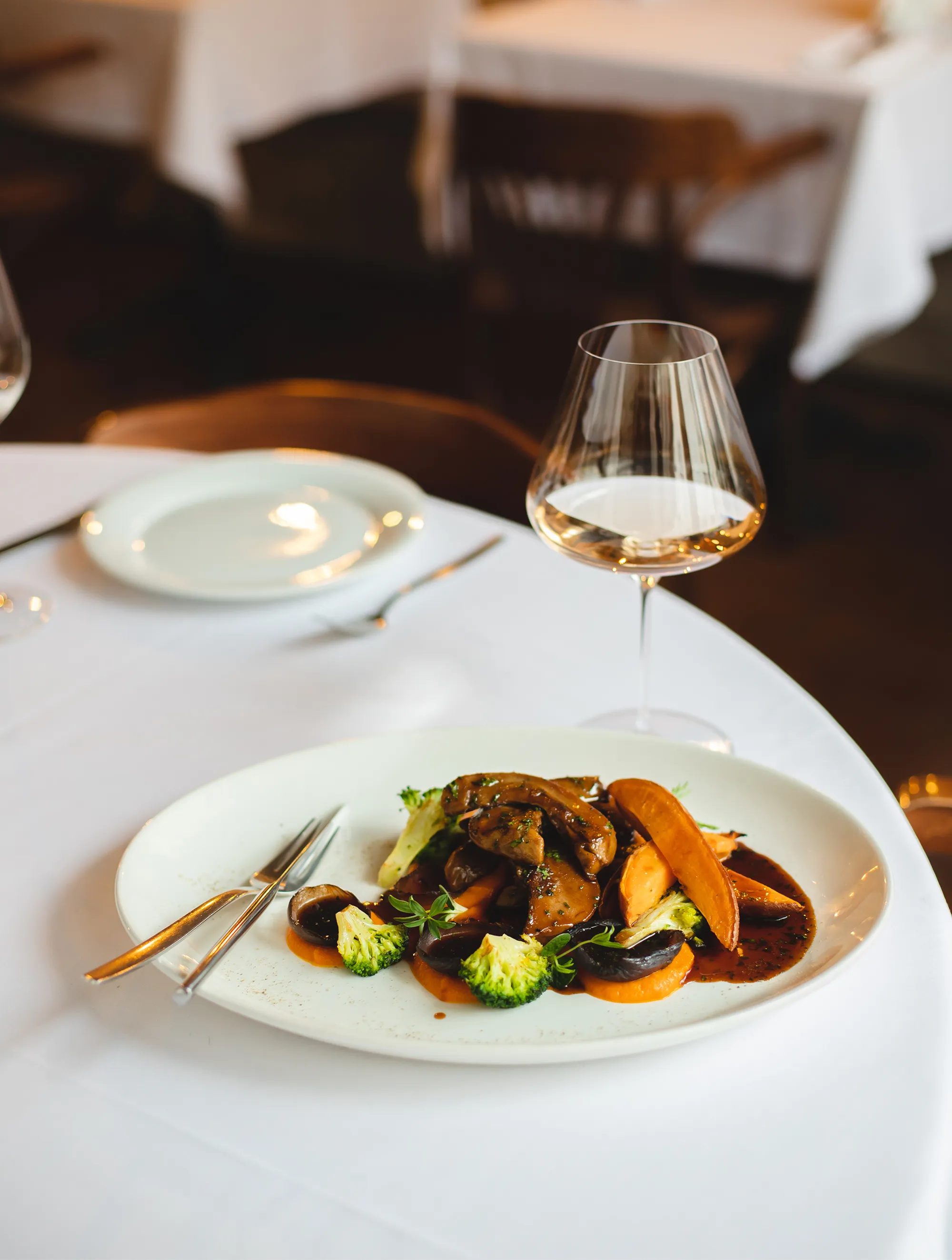 Gourmet plate with sautéed mushrooms, broccoli florets, sweet potato wedges, and sauce, accompanied by a glass of white wine on a white tablecloth.