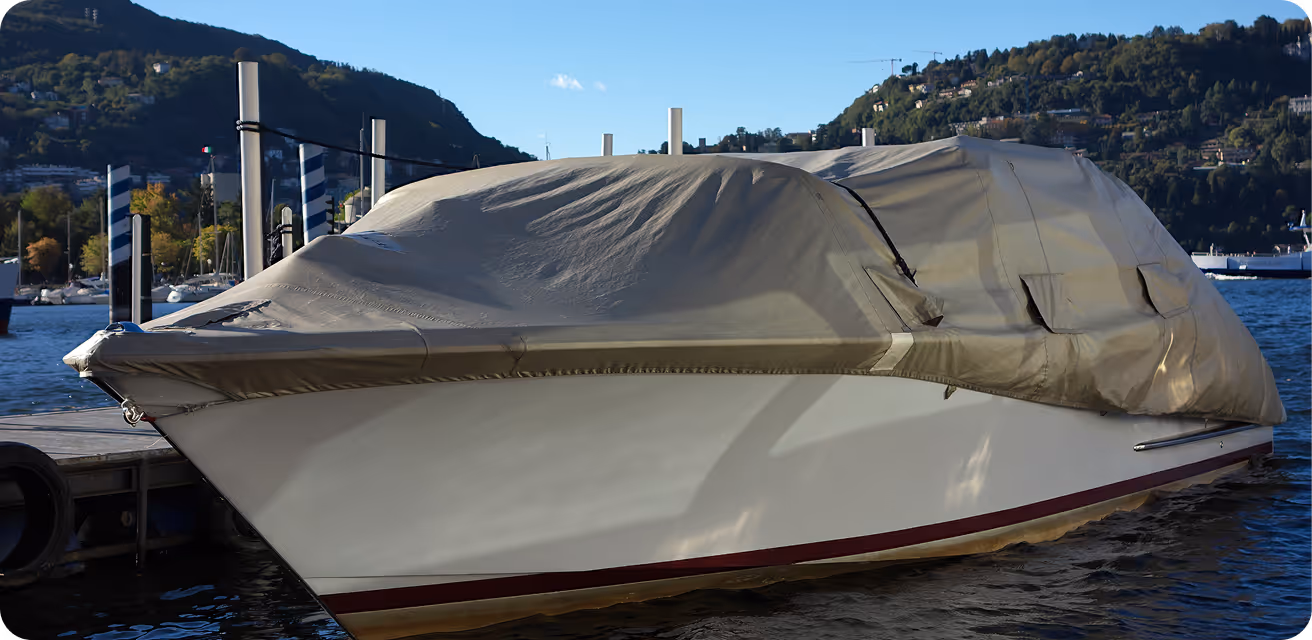 White motorboat covered with a fitted beige tarp docked on a lake with mountains and trees in the background.
