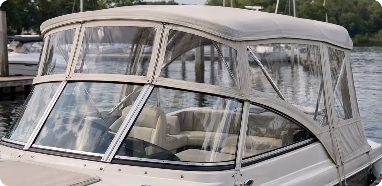 Close-up view of the front cabin of a white boat with clear plastic windows and beige canvas covers moored at a dock on a calm lake.