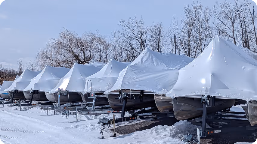 Row of boats covered with white protective tarps on trailers, parked on snowy ground with leafless trees in the background.