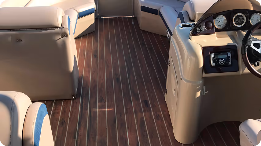 Interior view of a boat with wooden floor, beige cushioned seating, and control panel with steering wheel and gauges.