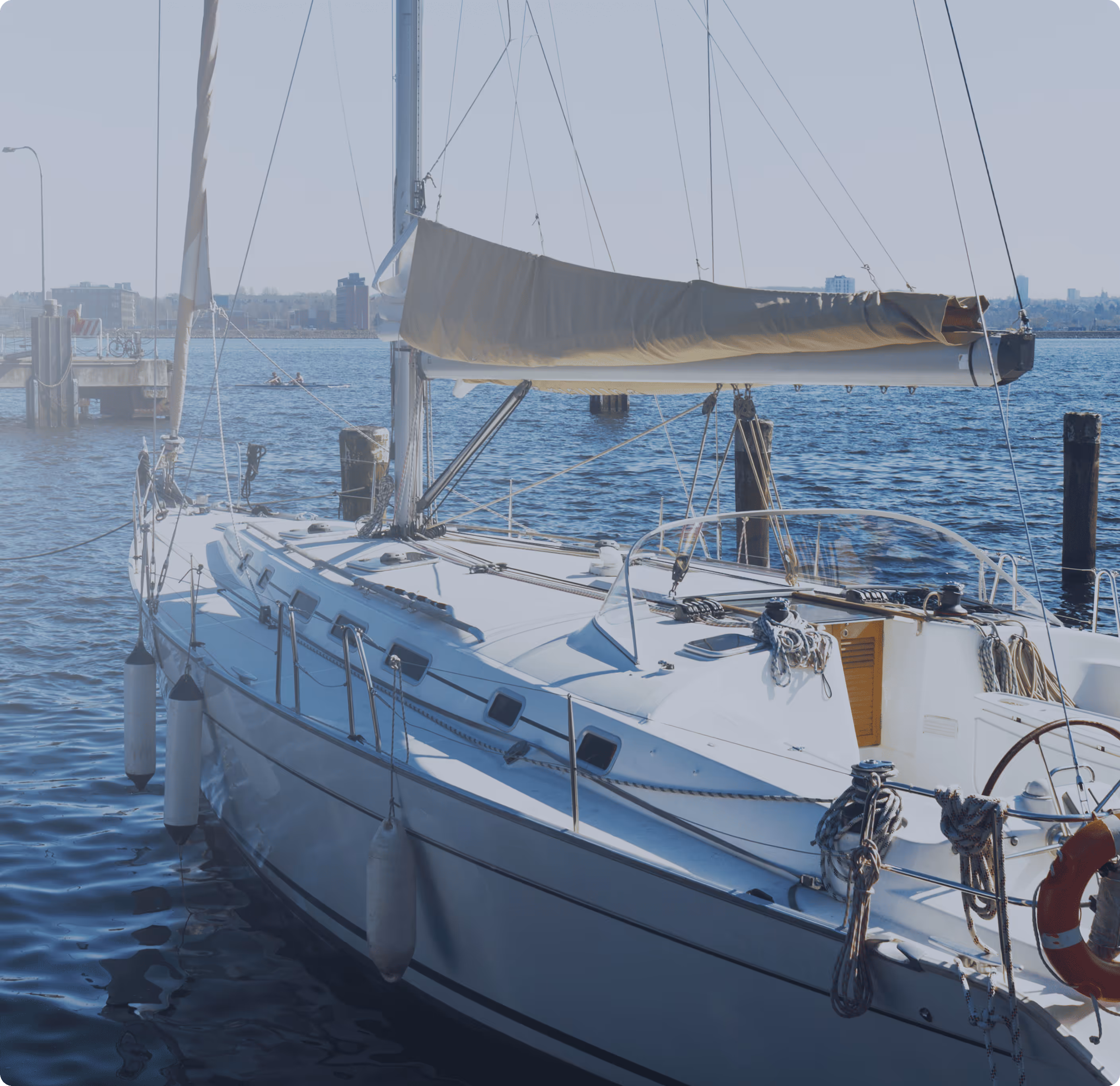 White sailboat docked on calm water with buildings and a pier in the background.