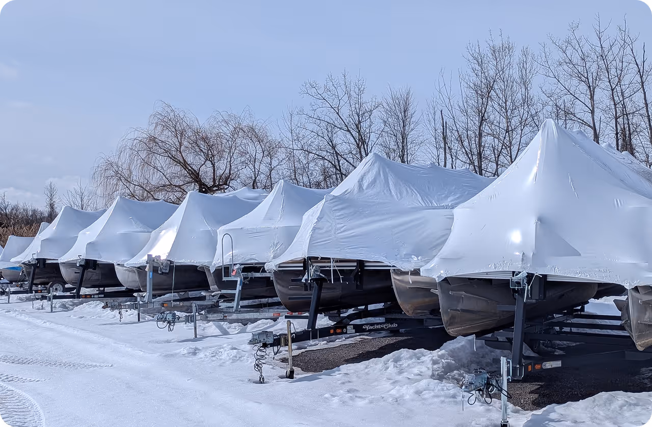 Row of boats covered with white protective shrink wrap on trailers in a snowy outdoor storage area with leafless trees in the background.
