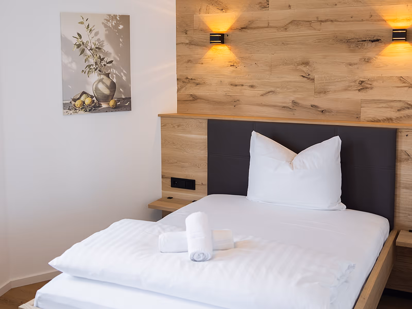 Ein ordentlich gemachtes Einzelbett mit weißer Bettwäsche, zwei Handtüchern darauf und Holzpaneelen an der Wand mit warmen Wandlampen.