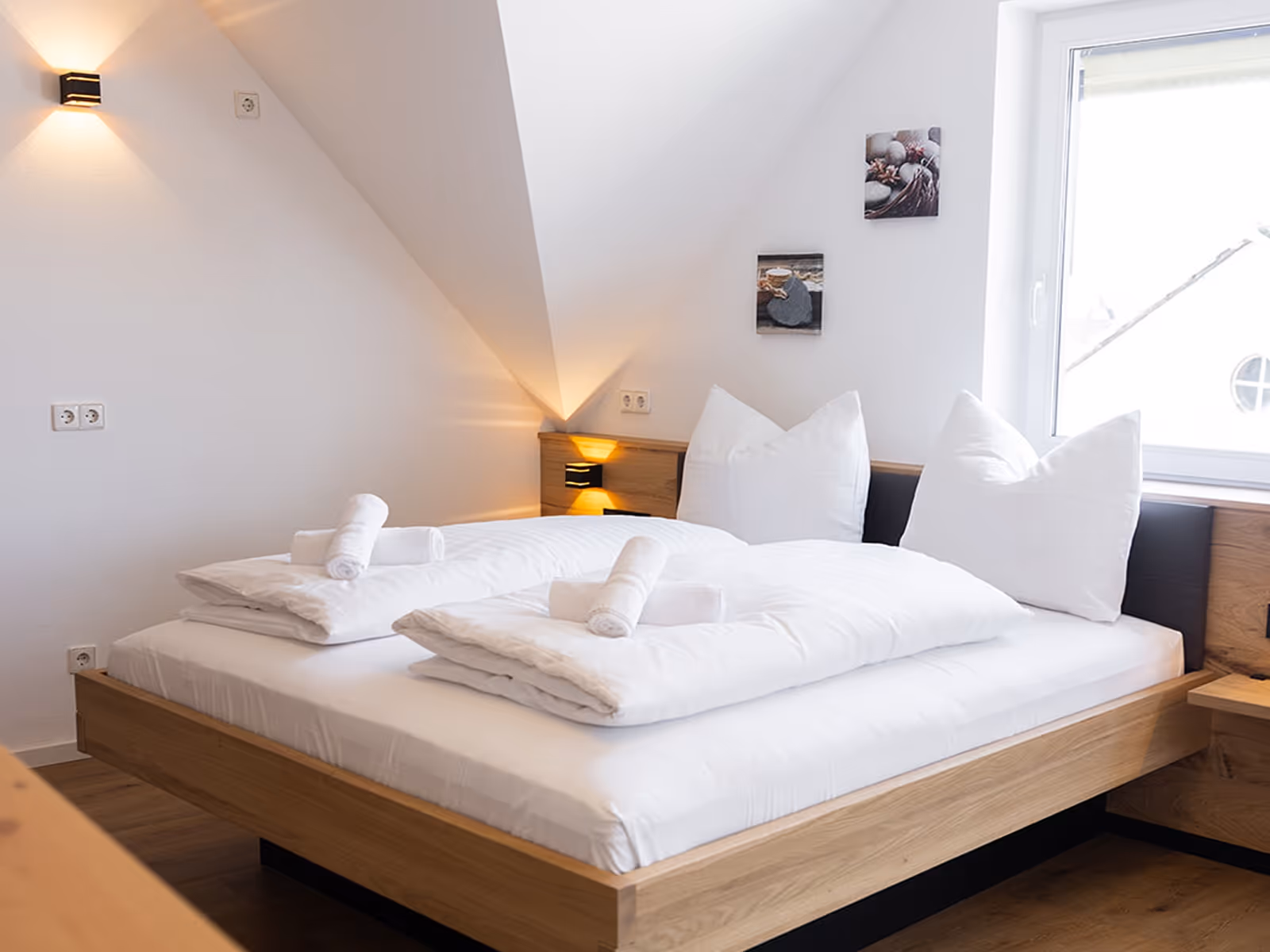 Hell eingerichtetes Schlafzimmer mit einem Doppelbett aus Holz, weißer Bettwäsche und Handtüchern, zwei Wandlampen und Fenster.