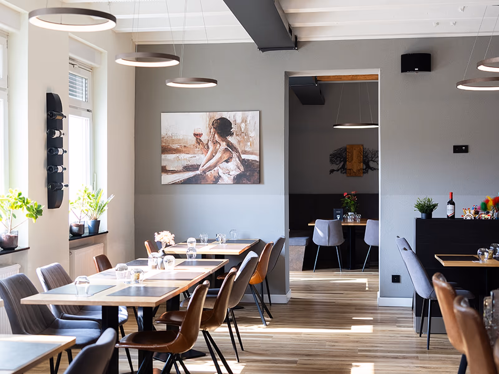 Modernes, helles Restaurant mit Holzboden, gemischten Stühlen und einem Gemälde einer Frau mit einem Weinglas an der grauen Wand.