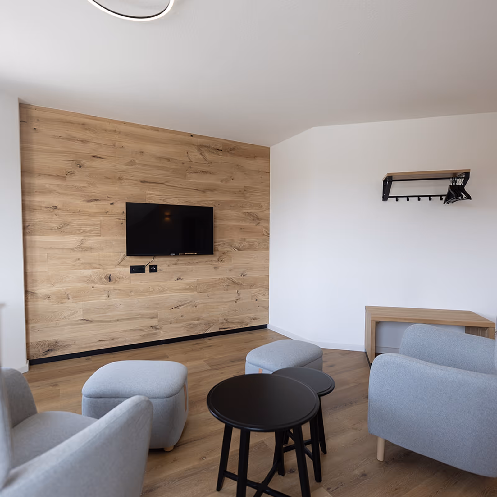 Modern Wohnzimmer mit hellgrauen Sesseln, schwarzen Beistelltischen und einem an einer Holzpaneelwand montierten Fernseher.