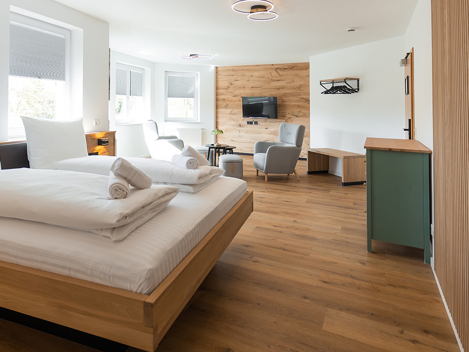 Modernes Hotelzimmer mit Holzfußboden, grauem Sessel und Hocker, zwei kleinen schwarzen Tischen, einem Bett mit weißen Bettwaren und einem grünen Schrank.