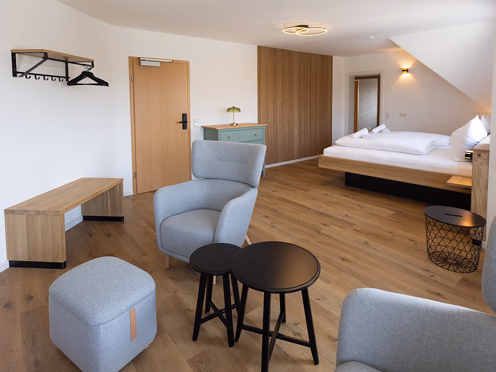 Modernes Hotelzimmer mit Holzfußboden, grauem Sessel und Hocker, zwei kleinen schwarzen Tischen, einem Bett mit weißen Bettwaren und einem grünen Schrank.