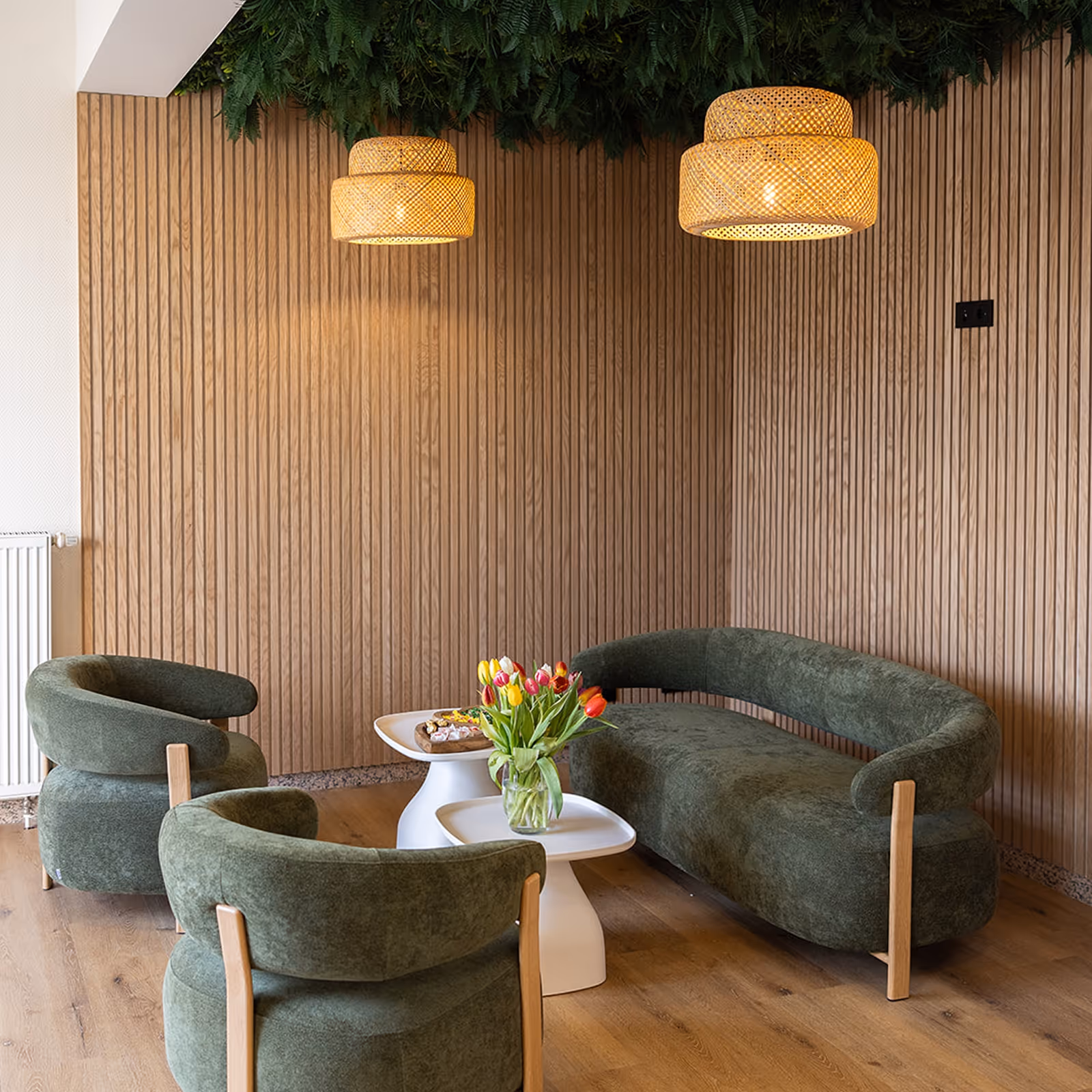 Wohnzimmer mit grüner Couch und Sesseln, weißen Beistelltischen, Tulpenstrauß und hängenden Rattanlampen.