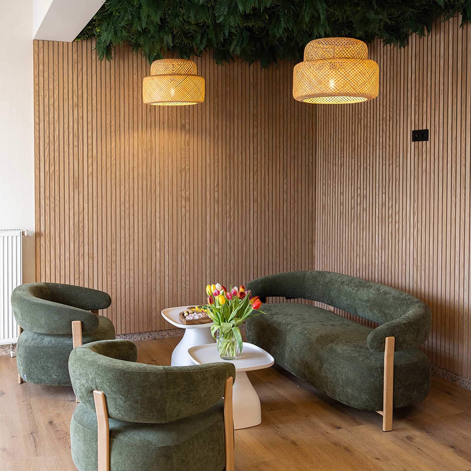 Wohnzimmer mit grüner Couch und Sesseln, weißen Beistelltischen, Tulpenstrauß und hängenden Rattanlampen.