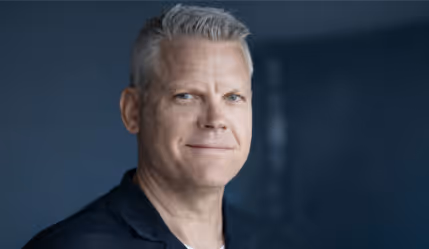 Porträtt av en mittålders man med gråsprängt hår, leende mot mörkblå bakgrund.