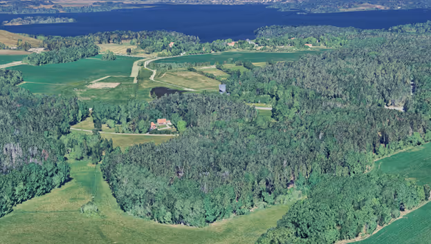 Flygvy över gröna jordbruksland och skogar med sjö och små röda hus.