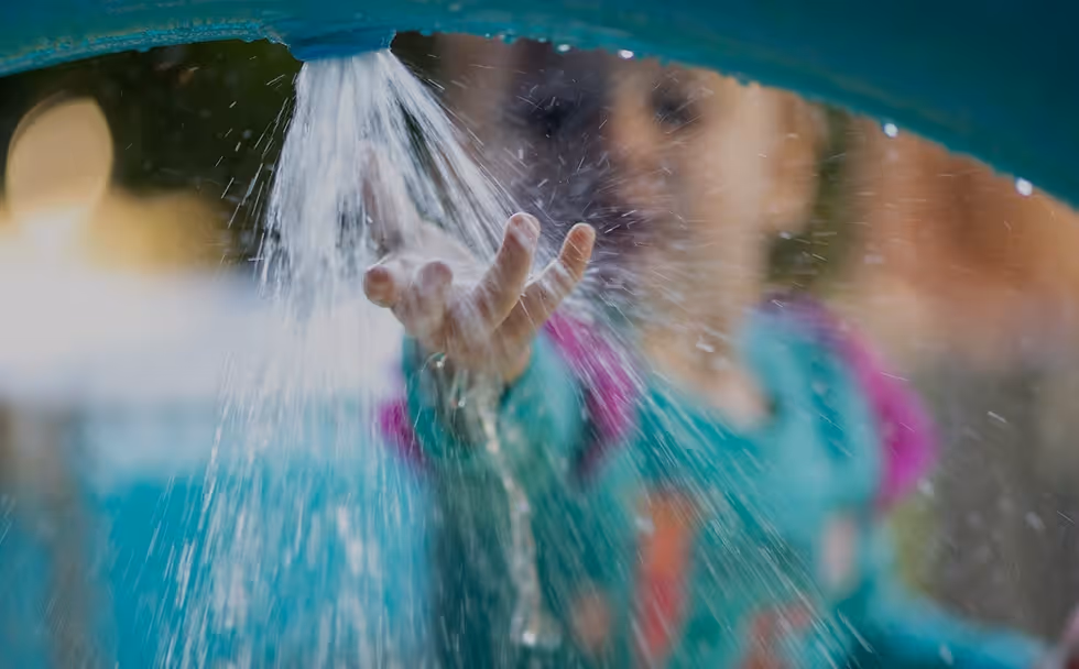 Kind spielt mit Wasser, das aus einer grünen Düse fließt und auf die ausgestreckte Hand trifft.