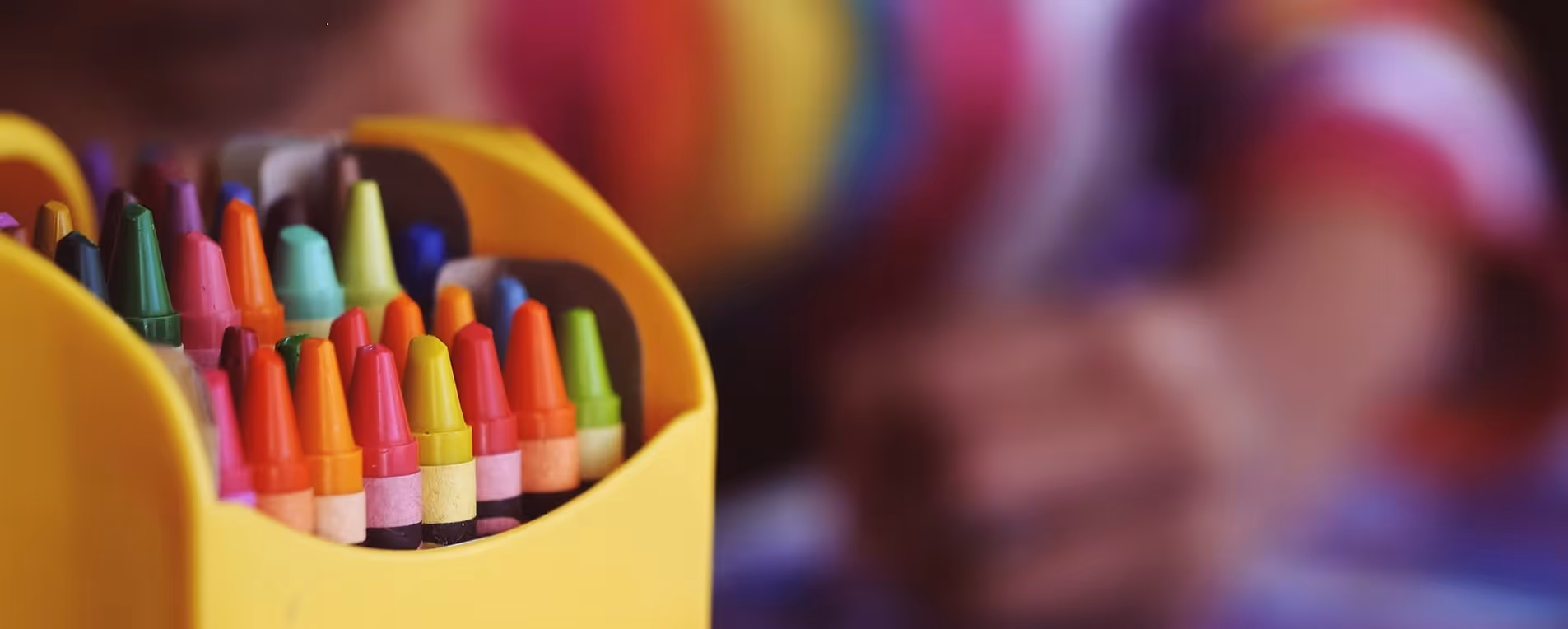 Eine Nahaufnahme von bunten Wachsmalstiften in einem gelben Behälter mit verschwommenen Händen im Hintergrund.