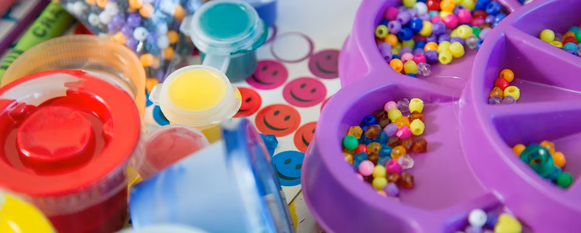 Nahaufnahme von bunter Bastelfarbe, Perlen und Aufklebern mit lachenden Gesichtern auf einem Basteltisch.