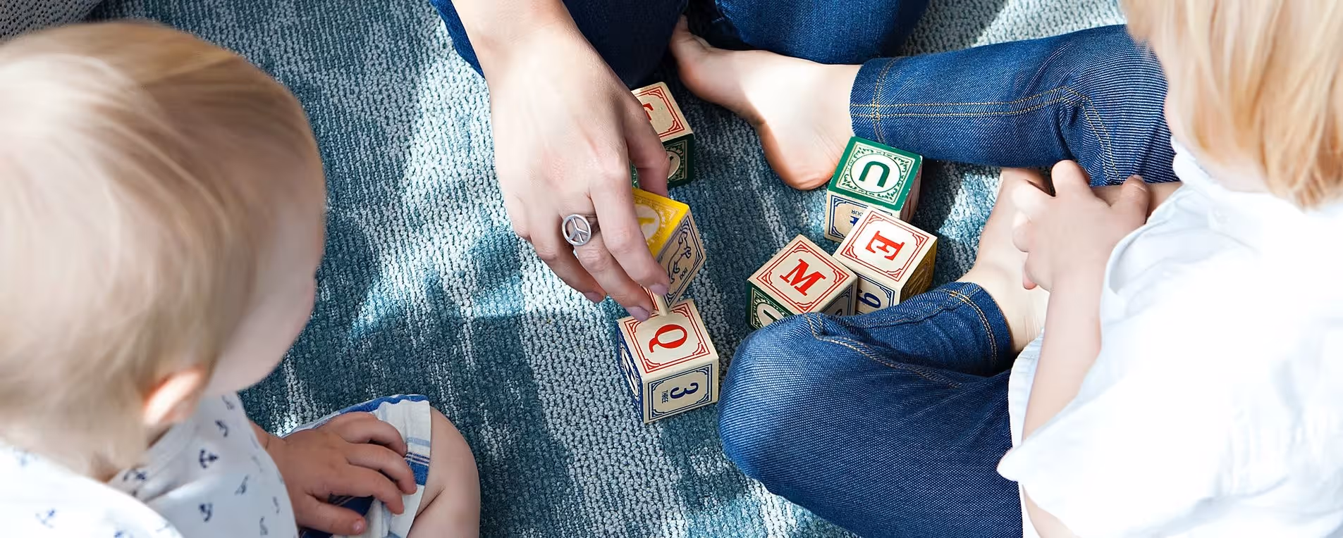 Zwei Kinder und eine erwachsene Person sitzen auf einem Teppich und spielen mit bunten Buchstabenwürfeln.