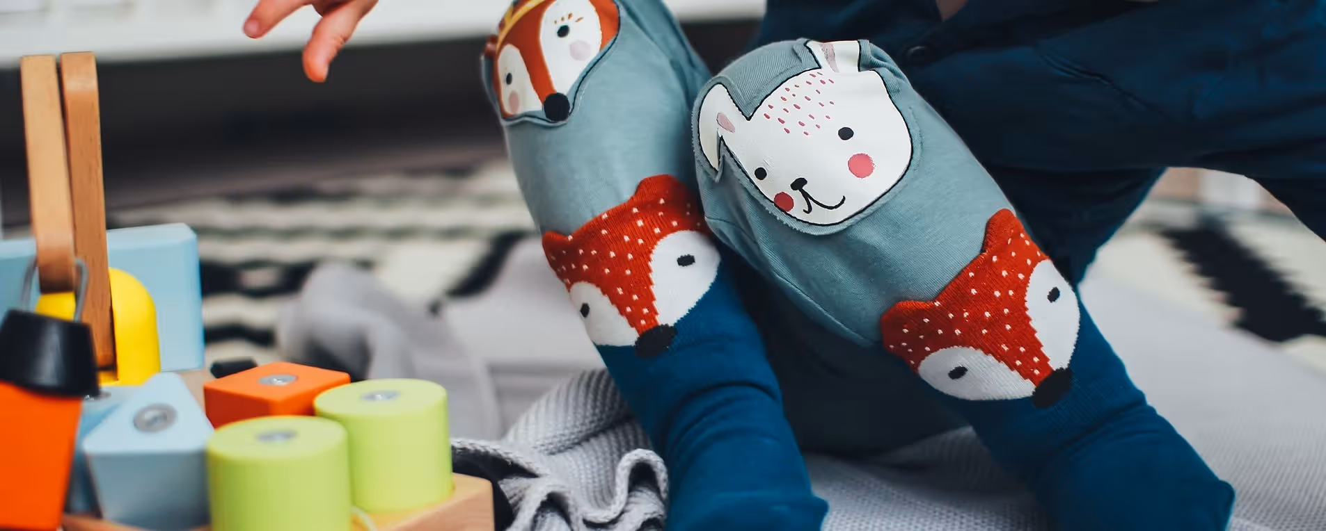 Kind trägt blaue Strumpfhose mit Fuchs- und Hundegesicht auf den Knien, sitzt auf dem Boden mit buntem Holzspielzeug.