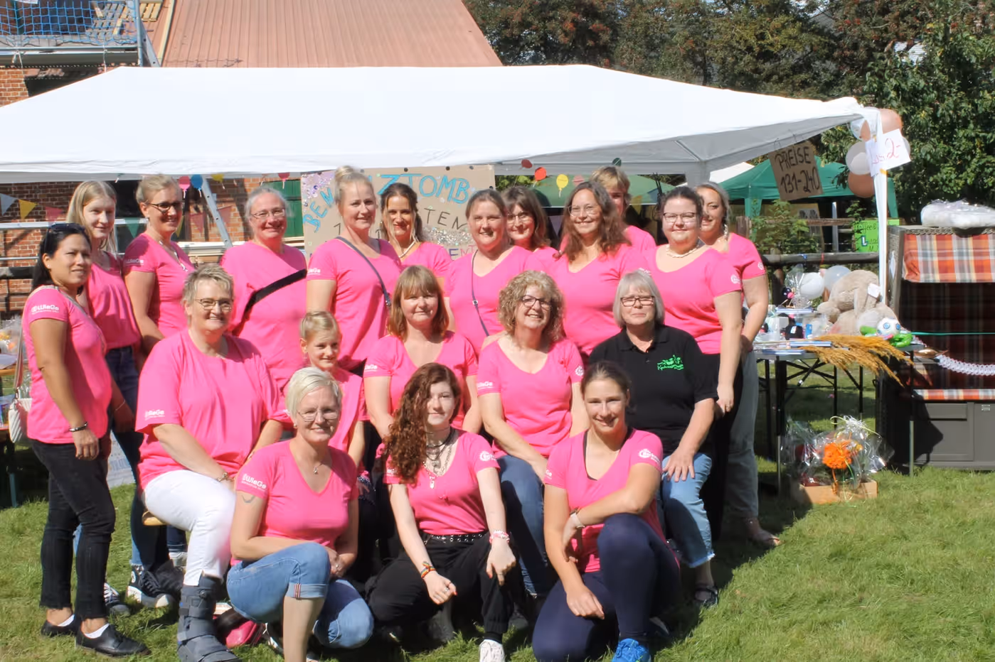 Gruppe von 20 Kindertagespflegepersonen in pinken T-Shirts posieren vor einem Pavillon bei einer Outdoor-Veranstaltung.