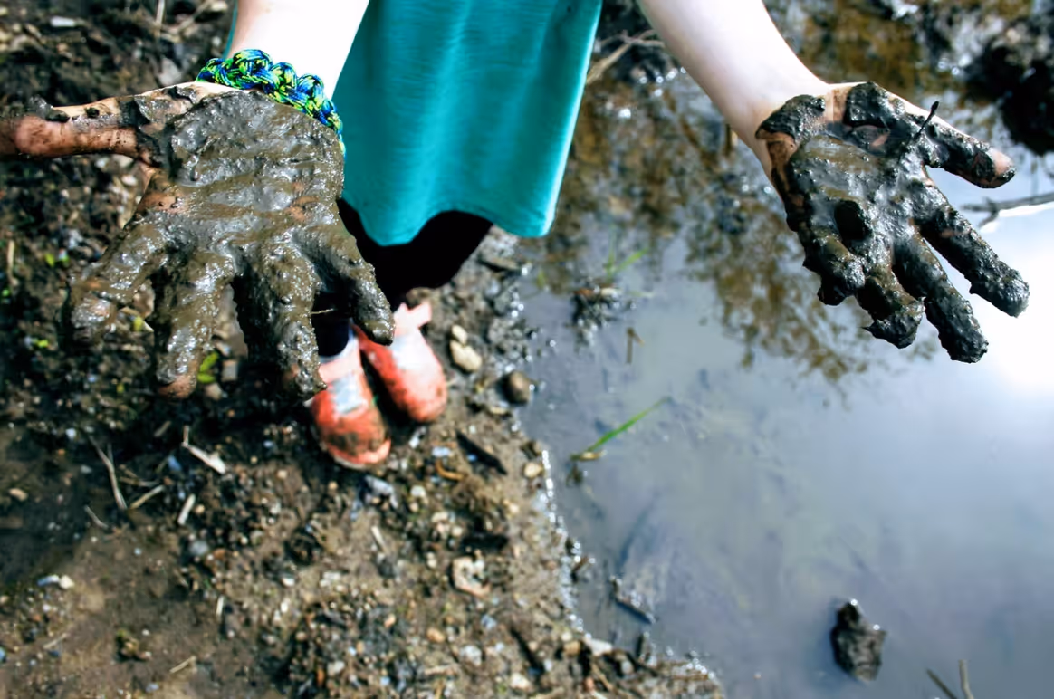 Kind hält schmutzige Hände  und steht mit roten Schuhen auf schlammigem Boden.